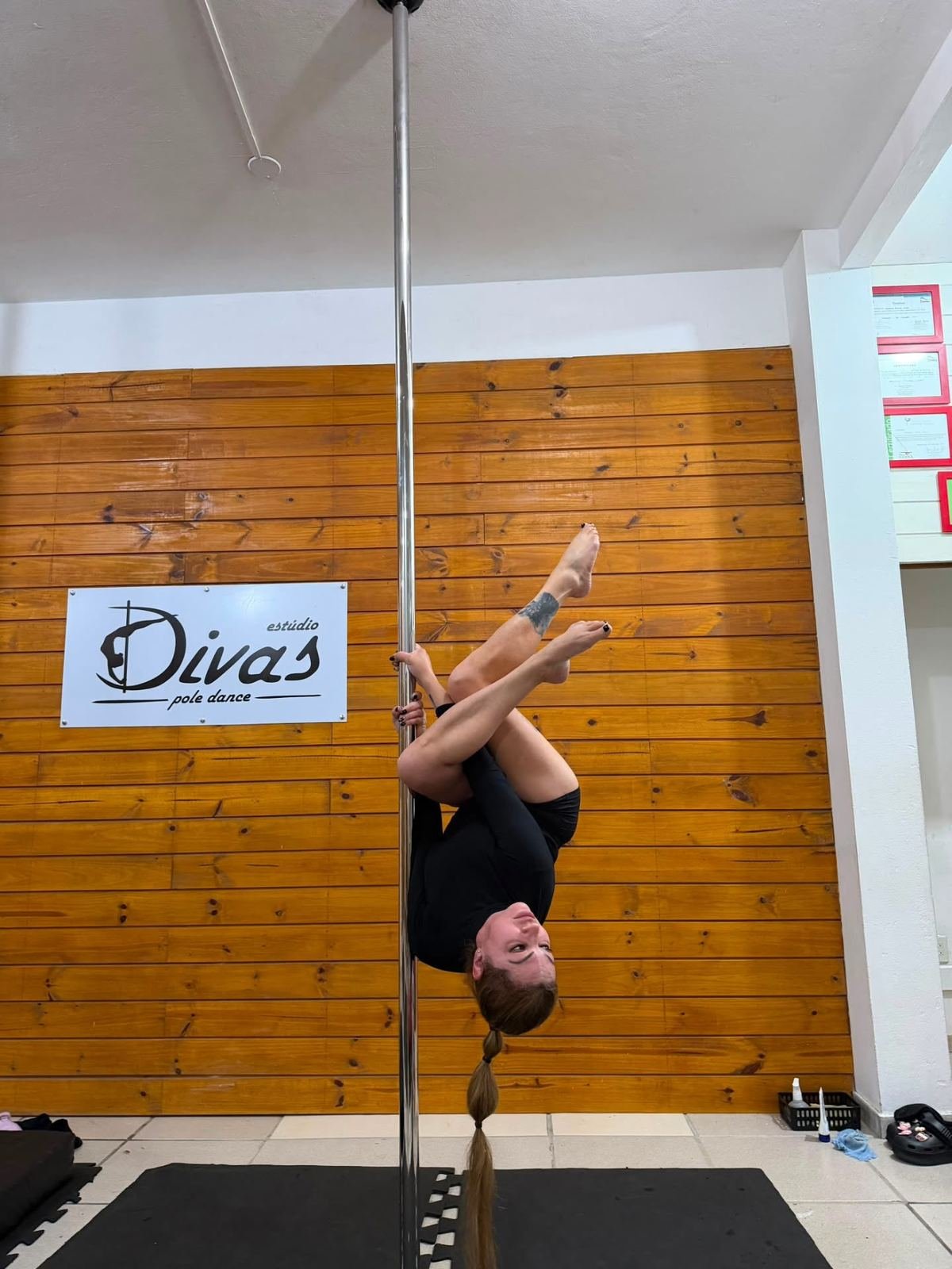Barra de pole inox com aluna suspensa, parede de madeira ao fundo, no Estúdio Divas