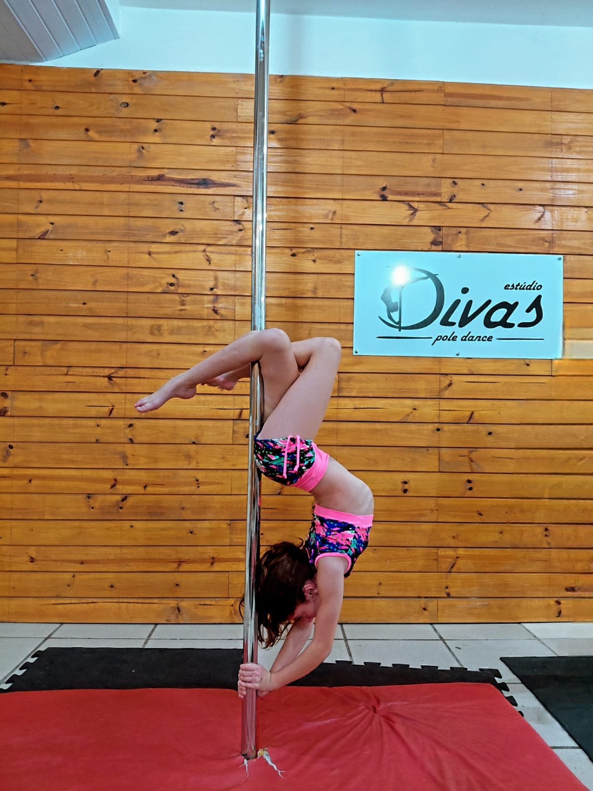 Criança praticando Pole Kids no Estúdio Divas, invertida na barra junto à placa da marca